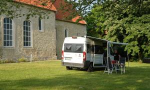 Vor einer Kirche steht ein Wohnmobil auf der grünen Wiese, davor steht ein Mann