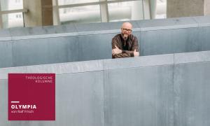 Ein Mann in Lederjacke steht hinter eine grauen Mauer, die Arme verschränkt, er lächelt