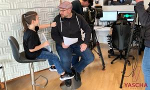 Ein junge und ein Mann sitzen auf Barhockern und sprechen miteinander, dahinter Leute mit Kameras