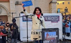 Eine Frau steht auf einer Bühne, umringt von Menschen. Sie hält ein Schild hoch: "Kein Platz für Nazis"
