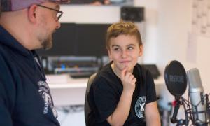 Ein Junge sitzt in einem Aufnahmestudio, vor ihm ein Mikrofon, neben ihm ein Mann
