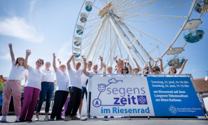 Auf dem Bild sieht man Menschen vor einem Riesenrad. Davor ist ein Banner mit der Aufschrift "Segenszeit im Riesenrad". 