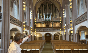 Pfarrer Ulrich Winkler in der Petrikirche Kulmbach mit der imposanten Orgel im Hintergrund, auf die der Pfarrer mit der Hand zeigt.