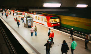 Menschen stehen vereinzelt an einer Ubahhaltestelle. Ein Zug fährt vorbei.
