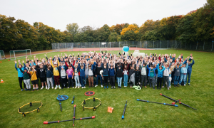 Gruppenbild von oben: Alle Jugendlichen stehen oder sitzen auf dem Rasen und heben gleichzeitig die Hände.