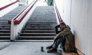 Ein Mann kauert unter einer Treppe am Bahnhof. Die Hände ins Gesicht abgestützt.
