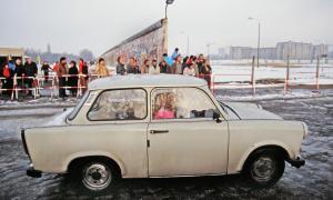 Ein Trabant fährt auf der Straße, dahinter Reste der Berliner Mauer