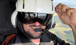 Ein Mann mit Flughelm auf, der in die Kamera lächelt. Im Hintergrund das innere des Flugzeugs, sowie die grüne Landschaft von oben. 