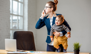 Eine Frau hält ein Baby im Arm, wärhend sie am Schreibtisch ist und telefoniert. Vor ihr ein aufgeklappter Laptop.