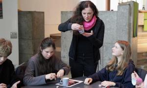 Eine Frau steht, Kinder sitzen an einem Tisch