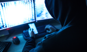 Person mit Hoodie über Kopf von hinten in einem dunklen Raum vor Handy und PC.