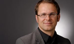 Ein Mann mit Brille, der in die Kamera lächelt. Grauer Hintergrund.