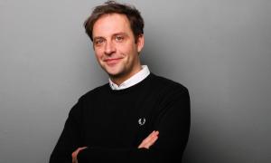 Nils Kumkar vor einer grauen Wand, die Arme verschränkt, in die Kamera lächelnd