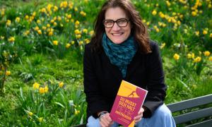 Melanie Dertinger hält ihr Buch in der Hand und kniet vor einem blühenden Wiese